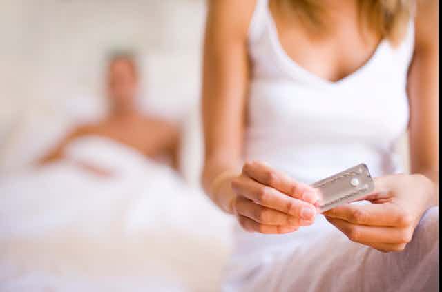 With her male partner lying in bed nearby, a woman opens a sealed package containing a pill.