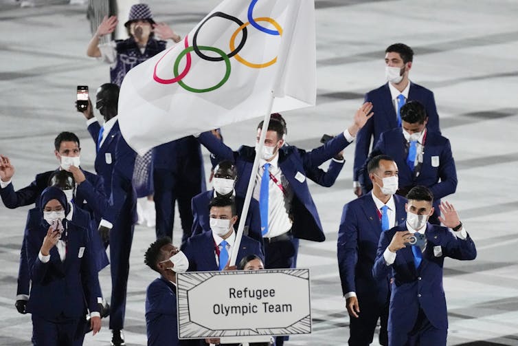 A group of people wearing dark blue suits wave behind a person carrying an Olympic flag and a sign that says 'Refugee Olympic Team'
