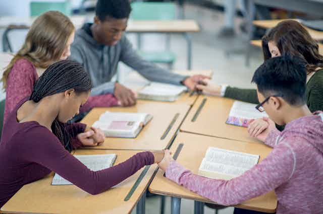 A diverse group of five teenagers hold hands, with heads bowed, in a classroom.