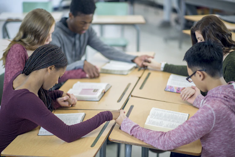 A diverse group of five teenagers hold hands, with heads bowed, in a classroom.