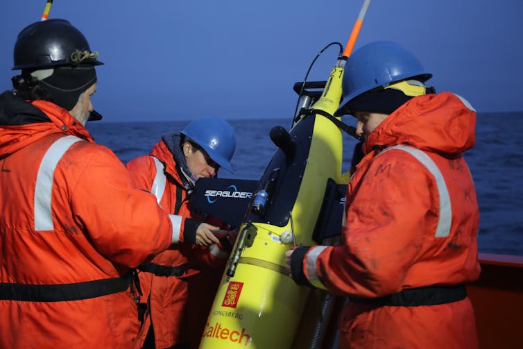Three people bundled up in winter gear work on a large seagoing drone.
