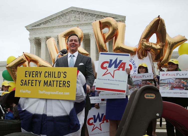 A man in a suit speaks in front of a crowd in front of the Supreme Court, with people holding up balloons that spell out 'fair play' behind him.