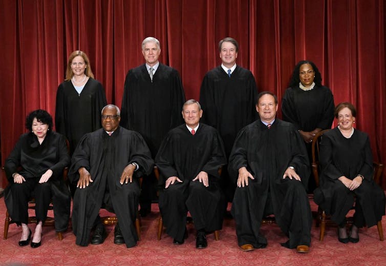 Two rows of people in black robes pose for a formal portrait in front of red velvety curtains.