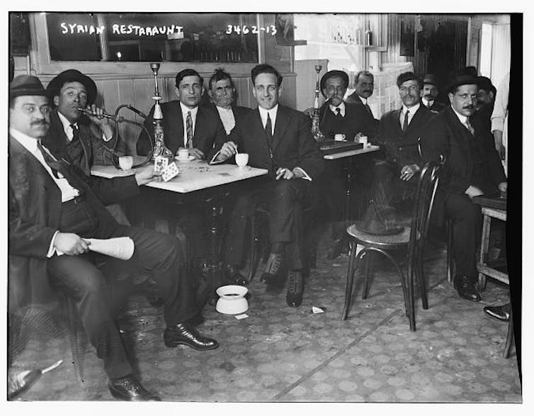A black and white photo shows a group of men in suits at cafe tables, looking at the camera.