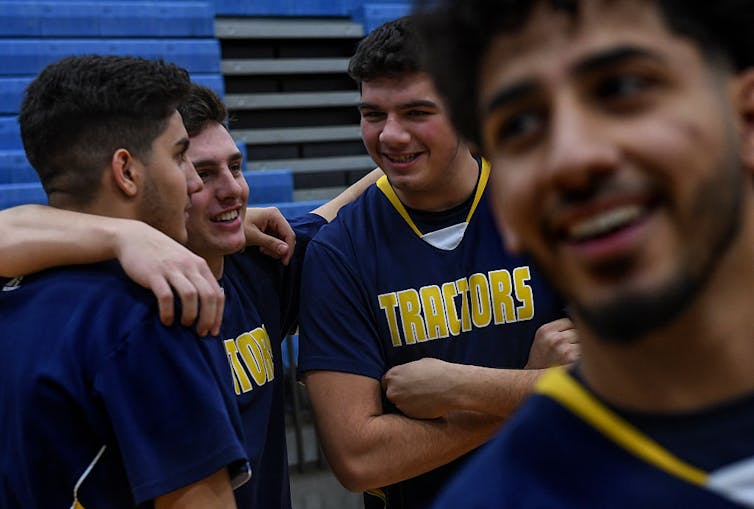 Three boys embrace, smiling, in basketball uniforms, while another laughs in front of them.