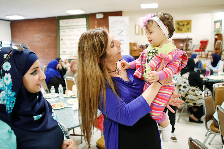 A woman in a headscarf looks on as another woman smiles and holds up a toddler in a pink striped outfit.