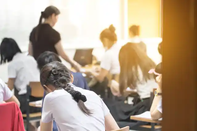 Students working, hunched over desks, with teacher in background.