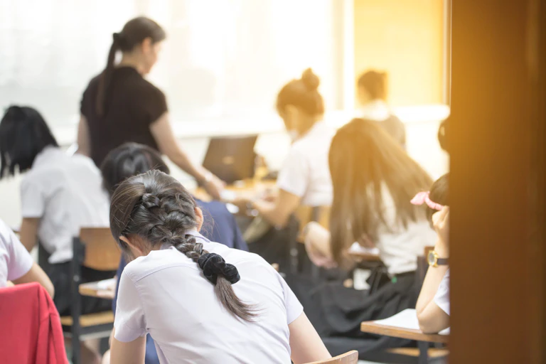 Students working, hunched over desks, with teacher in background.