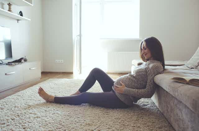 Mujer embarazada sentada en el suelo tocándose la barriga y sonriendo.