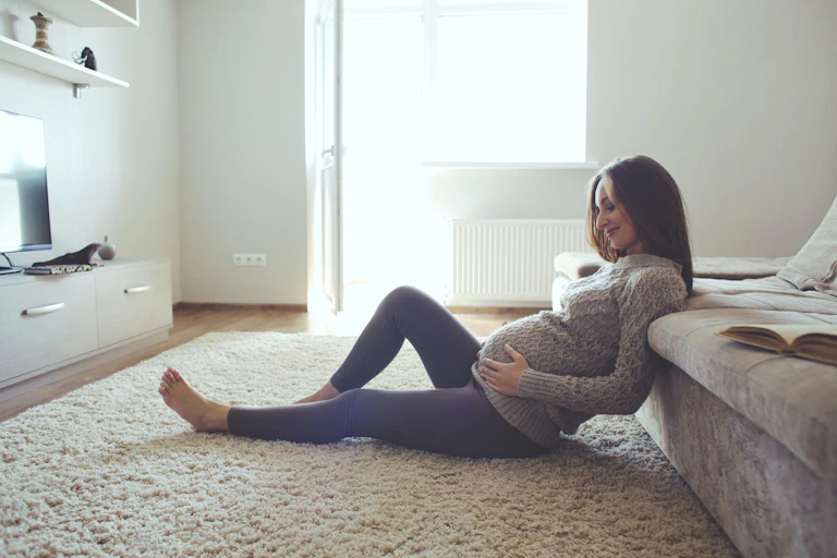 Mujer embarazada sentada en el suelo tocándose la barriga y sonriendo.