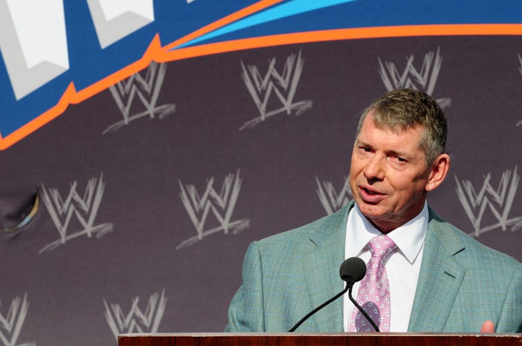 Vince McMahon speaking at a press conference wearing a grey suit.