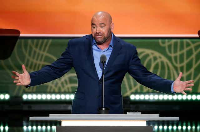 Dana White speaks at a conference, wearing a blue suit, his arms out stretched. 