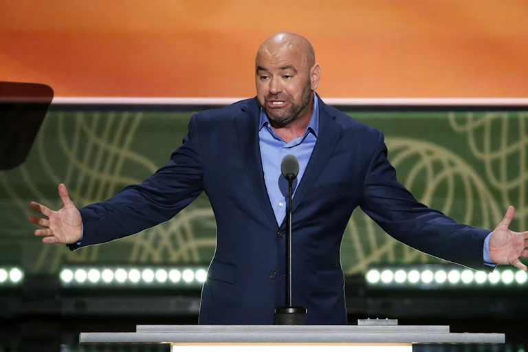 Dana White speaks at a conference, wearing a blue suit, his arms out stretched.