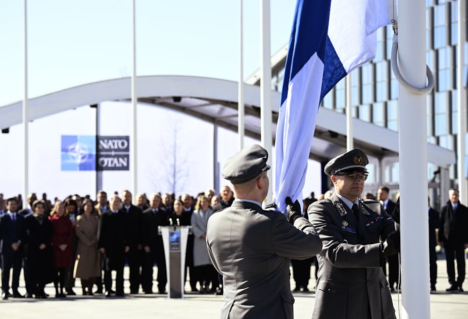 Deux militaires hissent le drapeau finlandais devant une assemblée de personnes derrière lesquelles on aperçoit un panneau officiel de l'OTAN