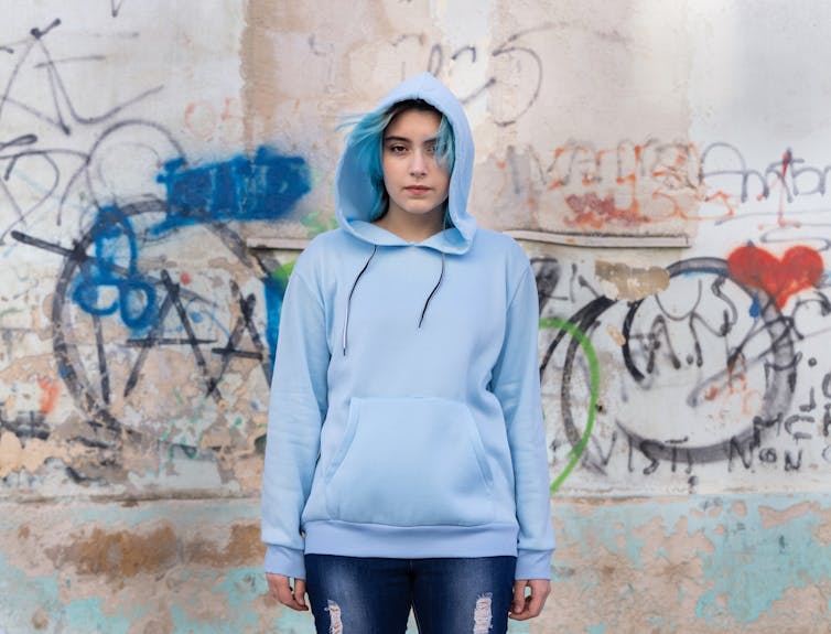 A girl with blue hair in a blue hoodie in front of a graffitied wall.