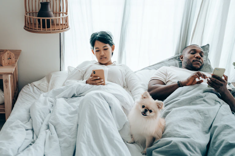 Multiracial couple surfing mobile phones lying in bed with dog