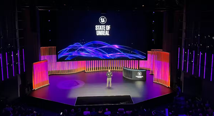 A man addressing an audience from the stage with a pink and blue lit background.