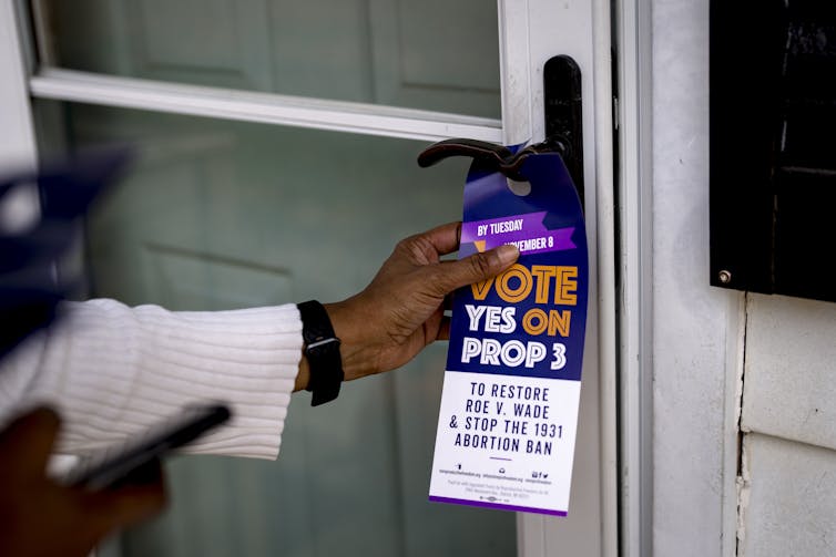 The arm of a Black woman, clad in a white sweater and black watch, is seen placing a tag that reads, 