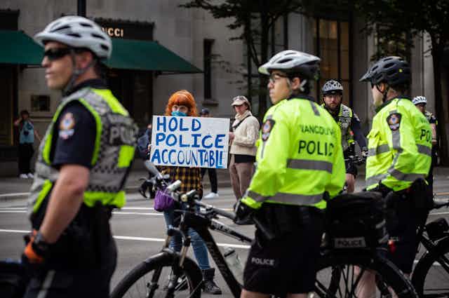 A woman standing near police officers holds a sign that reads: hold police accountable.