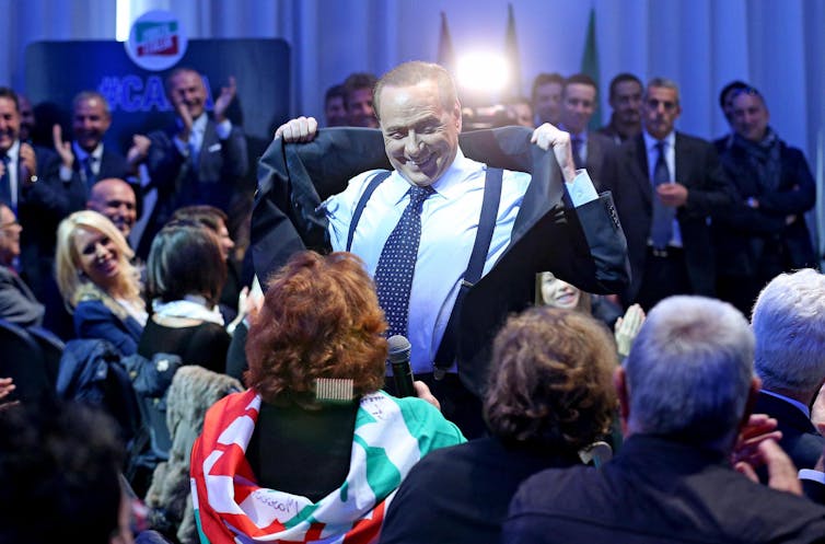 Silvio Berlusconi uśmiecha się, zdejmując kurtkę przed tłumem ludzi.