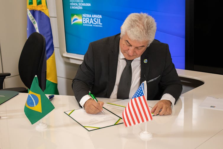 A man in a suit signing a document with a Brazilian and American flag on the desk.
