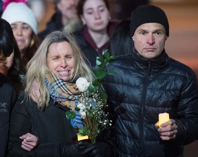 A weeping woman with blond hair walks among a crowd of people. A man in a black toque has his arm around her and a lit candle in his hand.