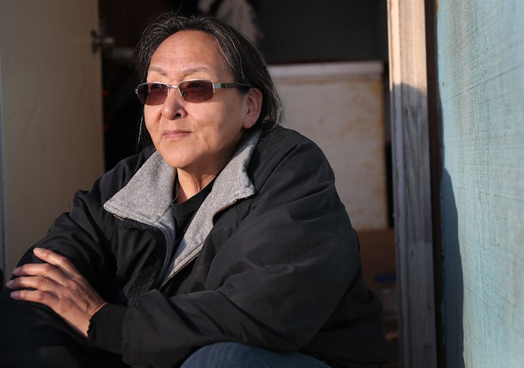 A woman wearing a fleece jacket and sunglasses sits in a doorway with her arms crossed.