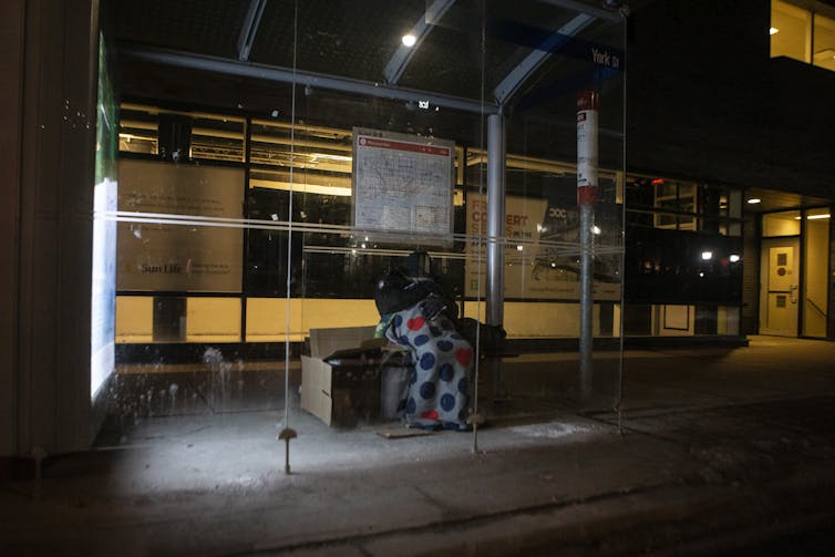 A homeless man sits in a bus shelter under a blanket.