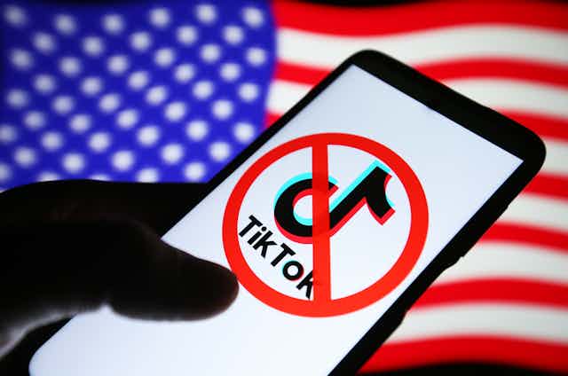 in the foreground a hand holds a smartphone displaying a logo with the circular 'no' symbol over it, in the background a U.S. flag 