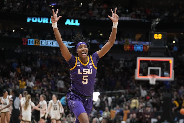 A female basketball player in a purple outfit raises her arms in celebration.