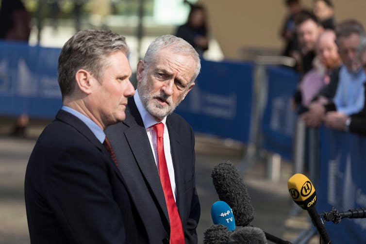 Keir Starmer talking into microphones while Jeremy Corbyn looks at him.