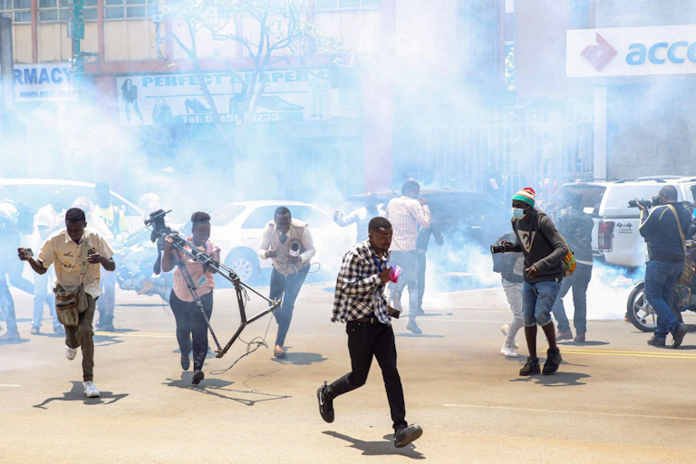 Men and women holding microphones and a camera stand running from a plume of smoke that is behind them