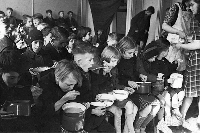 Niños comen de unos cazos sentados en filas.