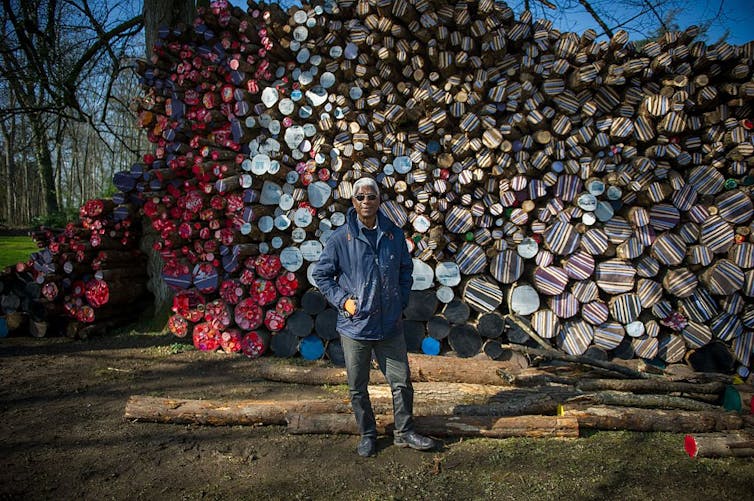 An elder man poses in front of a huge stack of wood, the ends of the logs covered in brightly coloured materials, forming a large painting-like work.