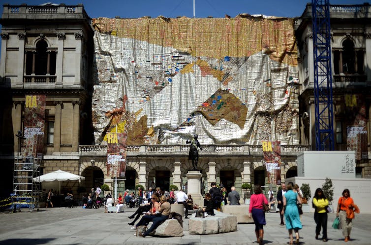 Hanging from a grand old building, a gigantic fabric shimmering in silver and gold covers a vast part of the building, people sit and stand observing it.