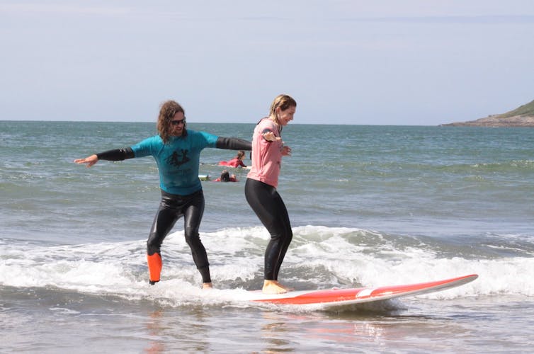 Two people stand on a surfboard and ride a small wave.