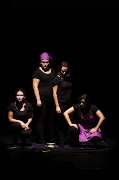 Varias actrices sobre el escenario vestidas de negro y con pañuelos violeta en la cabeza.