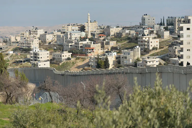 Several white buildings are seen in the distance, beyond a concrete wall and green, grassy areas.