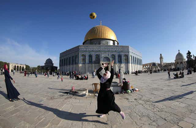 A girl wearing a dark dress and a grey head cover throws a ball in the air, in front of a large building that has a golden dome.