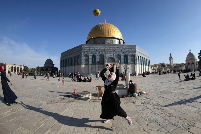 A girl wearing a dark dress and a grey head cover throws a ball in the air, in front of a large building that has a golden dome.