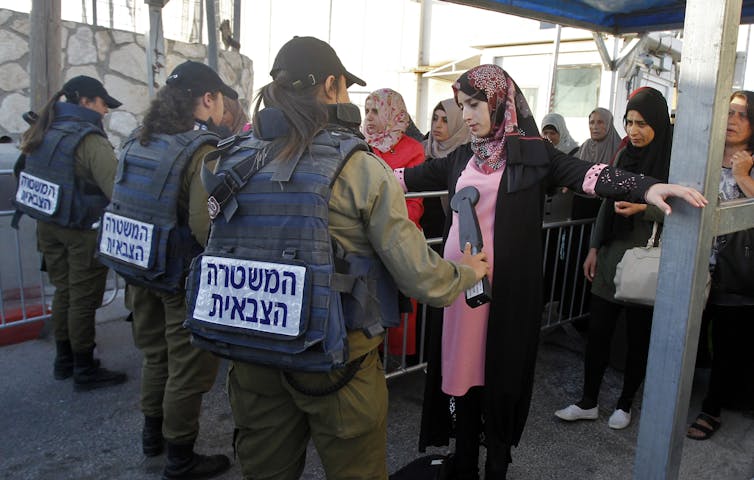 Mujeres con cobertores en la cabeza y vestidos largos se alinean y miran hacia las personas en verde militar, con escritura hebrea en la espalda.  Uno sostiene un detector de metales.