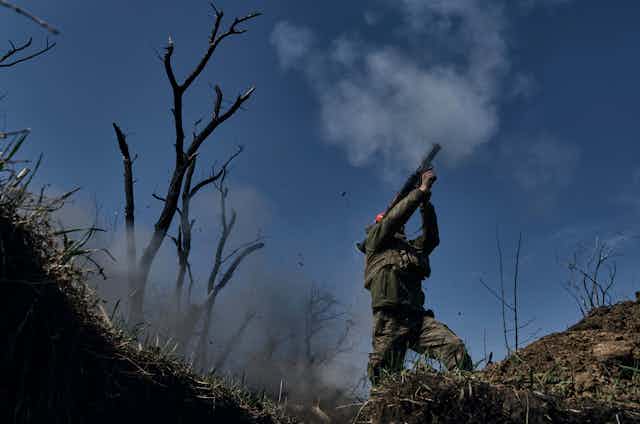 A man in battle fatigues fires a grenade launcher. Leafless trees are behind him.