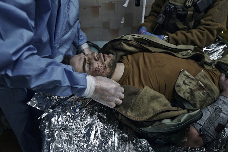 A young man with a dark beard lies on a gurney as a medic tends to him.