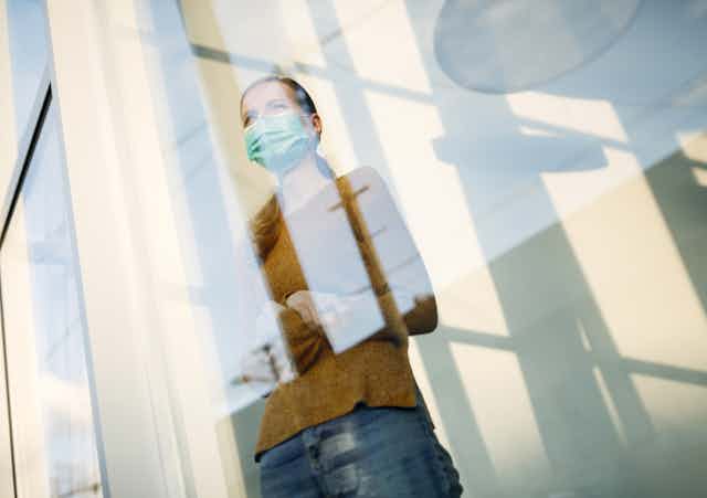 A woman in a face mask standing in front of a window looking out