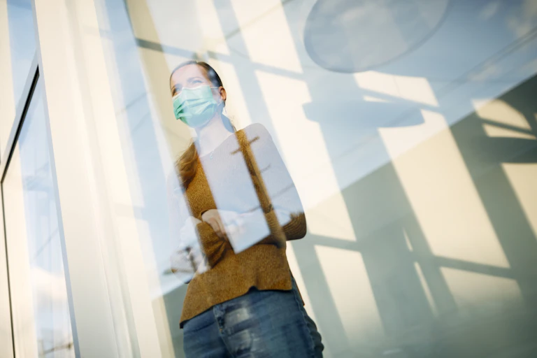 A woman in a face mask standing in front of a window looking out