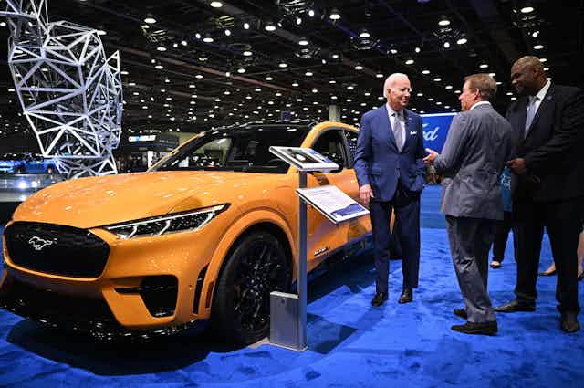 A bright yellow electric Ford Mustang car next to Biden and two other men.
