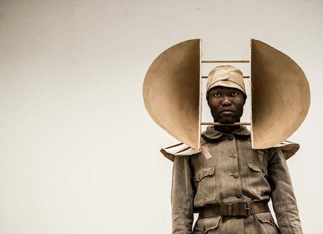 A stark image of a man in vintage army uniform. He stands looking straight ahead, on his head is mounted a large sculptural object with semi circular forms protruding from either side.