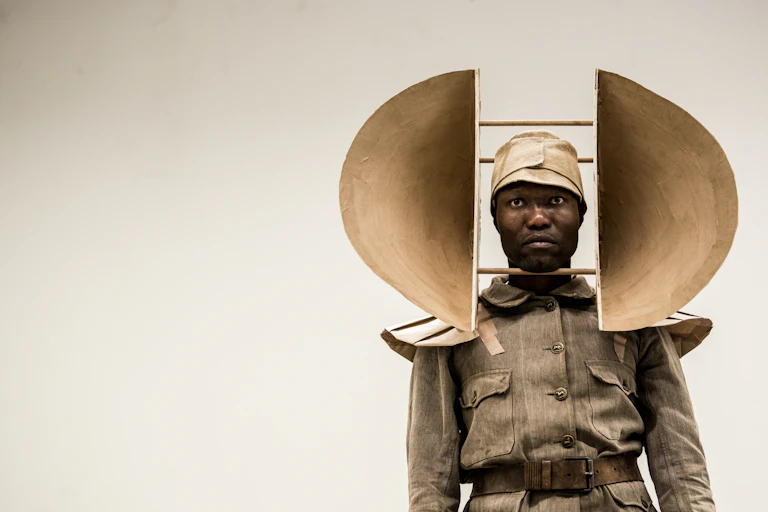 A stark image of a man in vintage army uniform. He stands looking straight ahead, on his head is mounted a large sculptural object with semi circular forms protruding from either side.