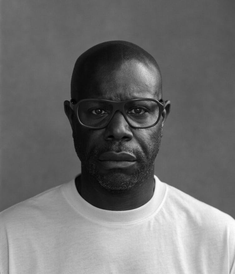 Steve McQueen looks straight to camera in a black and white photograph, wearing glasses and a white t-shirt.