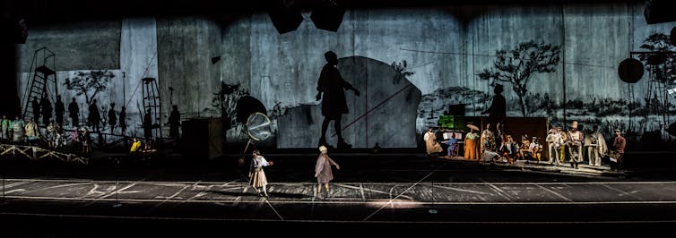 A wide angle photograph of a stage with figures and musicians, shadows cast onto a textured wall behind them.